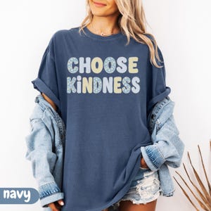 Puede incluir: Una camiseta de manga corta azul marino con el texto CHOOSE KINDNESS en una fuente redondeada y negrita. Las letras est&aacute;n rellenas con patrones alternos de flores azul claro, rayas amarillas y rayas azul claro sobre un fondo blanco.