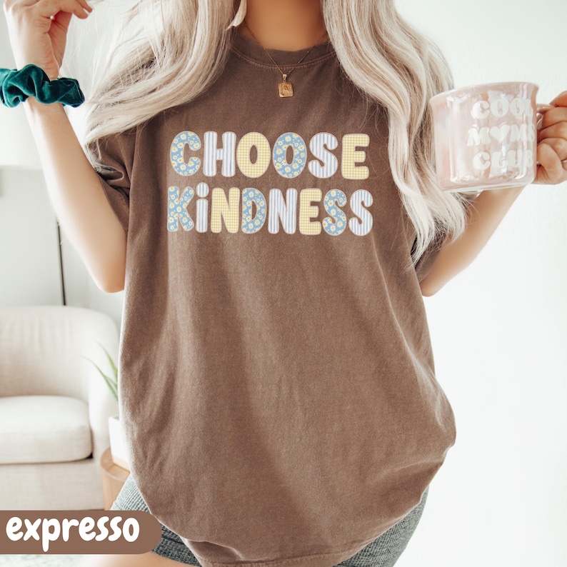 Puede incluir: Una camiseta marr&oacute;n de manga corta con el texto CHOOSE KINDNESS en una tipograf&iacute;a redondeada con patrones variados, incluyendo flores azules, cuadros vichy amarillos y rayas blancas. La prenda tiene cuello redondo y un corte holgado.