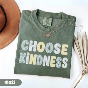 Puede incluir: Una camiseta de manga corta color verde musgo con cuello redondo y etiqueta Comfort Colors. El frente presenta el texto CHOOSE KINDNESS en letras grandes y redondeadas con patrones alternos de flores azules, cuadros vichy amarillos y rayas blancas.
