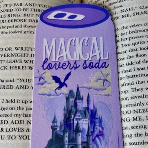 Puede incluir: Un marcapáginas morado con forma de lata de refresco que presenta una ilustración de un castillo, un dragón volador y estrellas. El texto MAGICAL lovers soda y support beverage aparece en una mezcla de fuentes serif y manuscritas.
