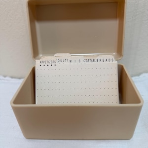 May include: A beige plastic recipe box with an open lid containing a set of off-white index cards. The visible tabbed dividers feature printed text including APPETIZERS, MISC, VEGETABLES, and BREADS, with a dotted grid pattern on the cards.