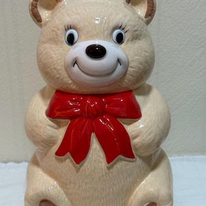 May include: A beige ceramic cookie jar shaped like a seated teddy bear. The bear features a glossy finish, a white muzzle, a black nose, painted blue eyes with long lashes, and a large, bright red bow tied around its neck. The paws have brown pads.