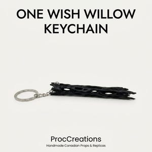 May include: A black One Wish Willow keychain featuring a cylindrical, porous, and textured wooden piece attached to a silver-colored metal split ring by a short chain. The text ONE WISH WILLOW KEYCHAIN and ProcCreations Handmade Canadian Props & Replicas appear.