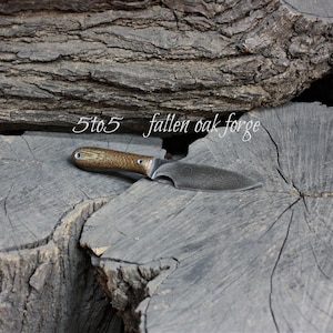 May include: A small, handcrafted knife with a textured, brown handle and a dark, curved blade. The knife rests on a weathered, gray wooden surface. The text "5to5 fallen oak forge" is above the knife.