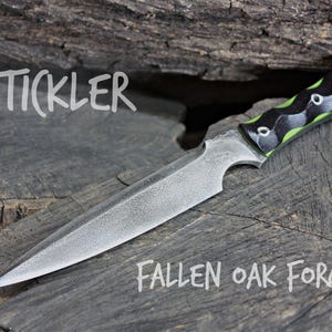 May include: A black and green handled knife with a textured blade. The knife is labeled "STICKLER" and "FALLEN OAK FORGE".