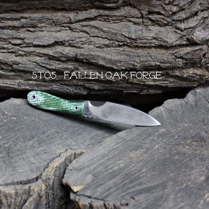 May include: A small, handcrafted knife with a green and white handle and a silver blade. The text "5T05 FALLEN OAK FORGE" is printed above the knife. The knife is displayed on a weathered, dark wood surface.