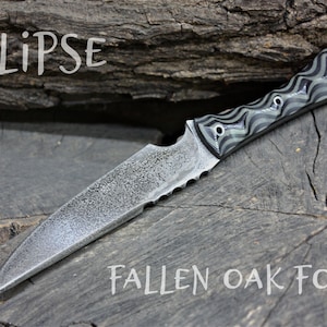 May include: A black and green patterned knife with a serrated blade and the text "ECLIPSE" and "FALLEN OAK FORGE" on a wooden surface.