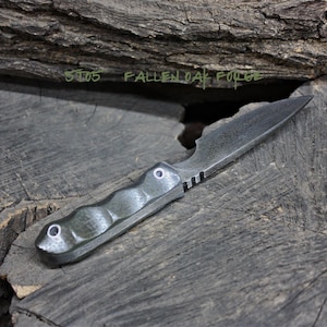 May include: A dark gray knife with a black handle and a textured surface. The knife has a small, pointed blade and a rounded handle. The knife is sitting on a piece of wood. The text "ST05 FALLEN OAK FORGE" is written on the wood.