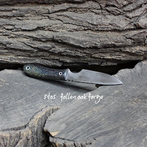 May include: A handcrafted knife with a textured, dark green and black handle and a silver blade. The knife rests on a weathered wooden surface, with the text "stos fallen oak forge" in white script.