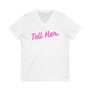 May include: A white short-sleeved t-shirt with a V-neckline. The center of the chest features the text "Tell Her." printed in a bright pink, cursive script font. The garment is plain white with short sleeves and a standard hemline.