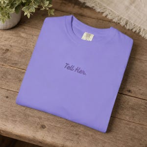 May include: A light purple short-sleeved t-shirt featuring the embroidered text "Tell Her." in a dark purple cursive font centred on the chest. The garment has a crew neckline and a visible brand label inside the collar area.