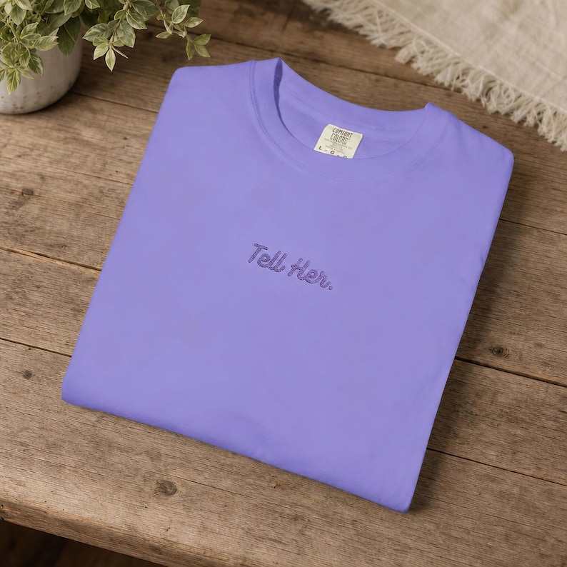 May include: A light purple short-sleeved t-shirt featuring the embroidered text "Tell Her." in a dark purple cursive font centred on the chest. The garment has a crew neckline and a visible brand label inside the collar area.