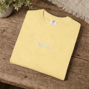 May include: A pale yellow short-sleeved t-shirt featuring the embroidered text "Tell Her." in a white cursive font centred on the chest. The garment includes a white interior neck label with the brand name Comfort Colors printed in black.