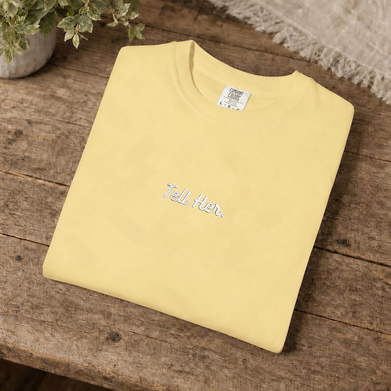 May include: A pale yellow short-sleeved t-shirt featuring the embroidered text "Tell Her." in a white cursive font centred on the chest. The garment includes a white interior neck label with the brand name Comfort Colors printed in black.