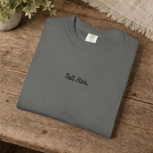 May include: A charcoal grey crewneck t-shirt featuring the embroidered black text "Tell Her." centred on the chest. The garment has a standard round neckline and a visible interior brand label at the collar, showcasing a simple design.