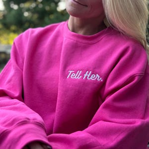 May include: A bright hot pink crewneck sweatshirt featuring the text Tell Her. embroidered in white cursive script on the upper left chest area. The garment has long sleeves with ribbed cuffs and a ribbed neckline, constructed from a solid fabric.
