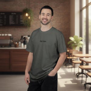 May include: A charcoal grey short-sleeve crew neck t-shirt featuring the text "Tell Her." printed in a small, black, cursive font on the upper left chest area. The garment is made of a solid-coloured fabric with a standard fit and hemline.