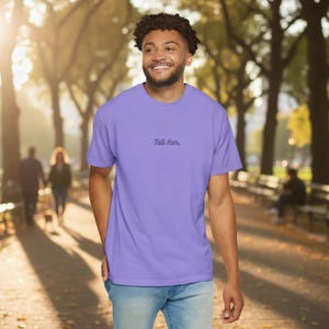 May include: A light purple short-sleeved t-shirt featuring the text "Tell Her." printed in a dark purple script font across the centre of the chest. The garment has a crew neckline and a standard fit, paired with light blue denim jeans.