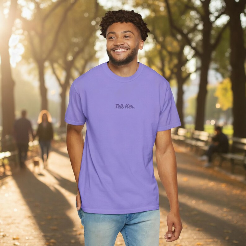 May include: A light purple short-sleeved t-shirt featuring the text "Tell Her." printed in a dark purple script font across the centre of the chest. The garment has a crew neckline and a standard fit, paired with light blue denim jeans.