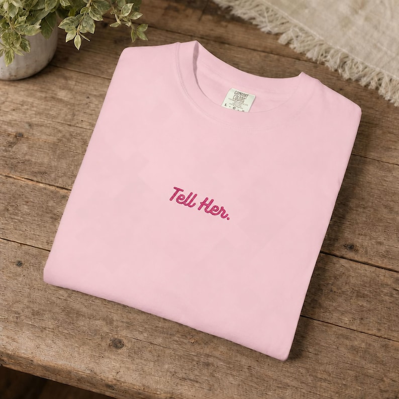 May include: A light pink crew neck t-shirt featuring the text Tell Her. embroidered in dark pink cursive script on the centre chest. The shirt has a visible interior neck label with the brand name Comfort Colors and a size L indicator.
