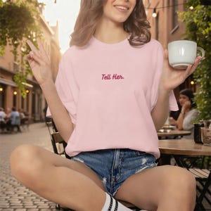 May include: A light pink short-sleeved t-shirt featuring the text "Tell Her." embroidered in a dark red cursive font on the centre chest. The garment has a crew neckline and a relaxed fit, paired with blue denim shorts with frayed edges.