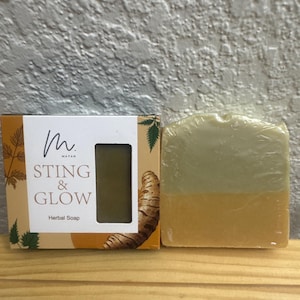 May include: A rectangular bar of herbal soap wrapped in clear plastic, featuring a two-tone design with a light green top and a golden yellow bottom. Beside it is a cardboard box labelled M. MAYAN STING & GLOW Herbal Soap with leaf graphics.