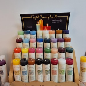 Puede incluir: Un expositor de madera contiene numerosas velas cilíndricas de varios colores, cada una con una etiqueta con texto y un gráfico. El letrero en la parte superior dice Crystal Journey Candles, A Collection Of Unique Candles.