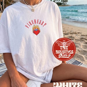 May include: A white short-sleeved t-shirt featuring a red heart graphic surrounded by blue and orange flames, with the text FIREHEART arched above in red lettering. The shirt is worn with a layered necklace featuring a small butterfly pendant.