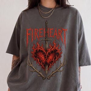 May include: A charcoal grey short-sleeve t-shirt featuring a graphic print of a red heart engulfed in flames, pierced by a vertical sword, and flanked by two deer antlers. The text FIREHEART is printed in a stylised red font above the heart.