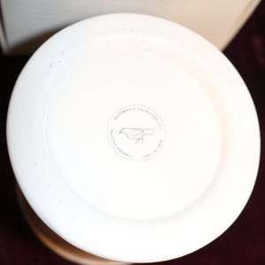 Pu&ograve; includere: Un vaso in ceramica bianca, visto dall'alto, con un bordo ampio e piatto. Sulla base &egrave; visibile un emblema circolare con l'illustrazione di un uccello. Il vaso ha una finitura liscia e lucida e un colore beige chiaro.