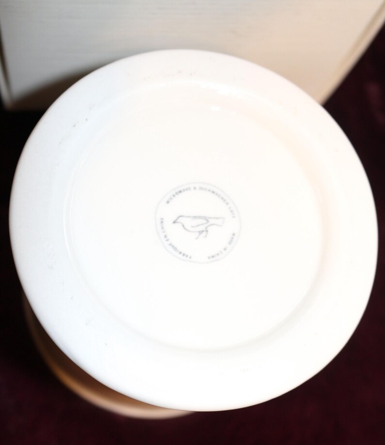 Pu&ograve; includere: Un vaso in ceramica bianca, visto dall'alto, con un bordo ampio e piatto. Sulla base &egrave; visibile un emblema circolare con l'illustrazione di un uccello. Il vaso ha una finitura liscia e lucida e un colore beige chiaro.