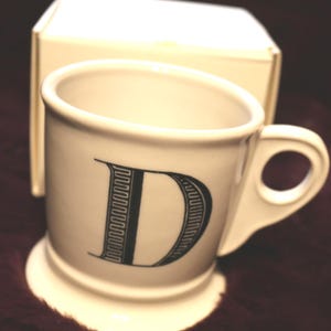 Pu&ograve; includere: Una tazza in ceramica bianca con la lettera "D" nera sul davanti. La tazza ha un piccolo manico e una base a piedistallo. Sullo sfondo c'&egrave; una scatola color crema.
