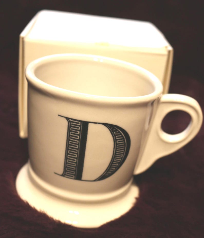 Pu&ograve; includere: Una tazza in ceramica bianca con la lettera "D" nera sul davanti. La tazza ha un piccolo manico e una base a piedistallo. Sullo sfondo c'&egrave; una scatola color crema.