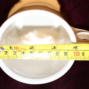 Pu&ograve; includere: Una tazza in ceramica bianca con manico marrone e una striscia marrone vicino alla parte superiore. L'interno &egrave; bianco. L'apertura della tazza misura circa 10 cm di diametro, come mostrato da un metro a nastro giallo.