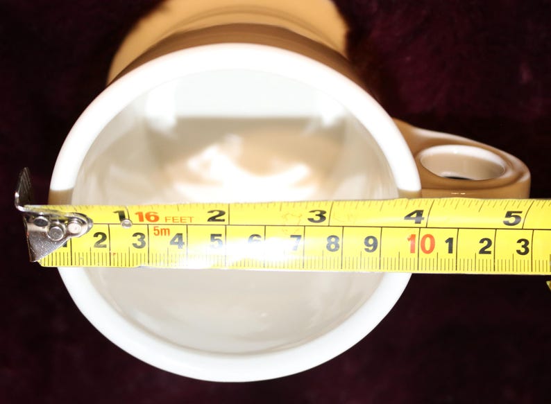 Pu&ograve; includere: Una tazza in ceramica bianca con manico marrone e una striscia marrone vicino alla parte superiore. L'interno &egrave; bianco. L'apertura della tazza misura circa 10 cm di diametro, come mostrato da un metro a nastro giallo.