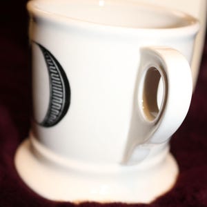 Pu&ograve; includere: Tazza in ceramica bianca con manico curvo e base larga. La tazza presenta un disegno in bianco e nero sul lato. La tazza &egrave; appoggiata su una superficie rosso scuro.