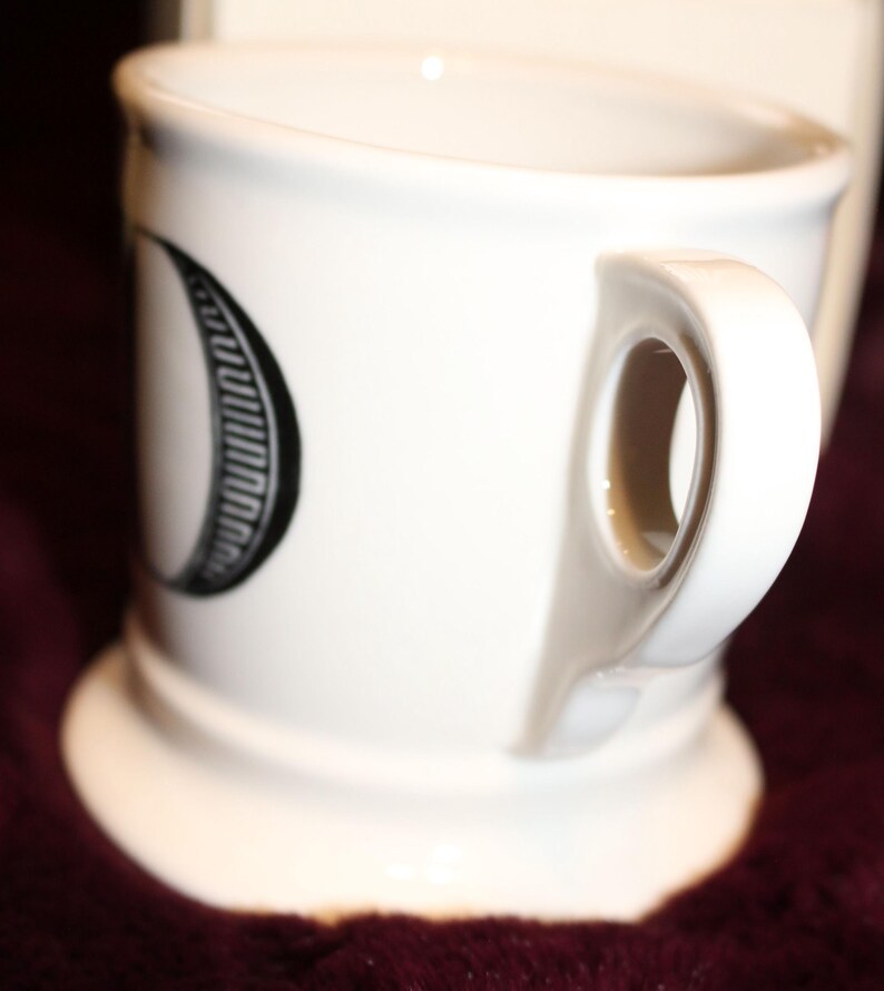 Pu&ograve; includere: Tazza in ceramica bianca con manico curvo e base larga. La tazza presenta un disegno in bianco e nero sul lato. La tazza &egrave; appoggiata su una superficie rosso scuro.