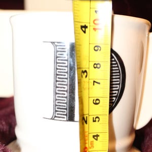 Pu&ograve; includere: Tazza in ceramica bianca con un design a lettera "I" nera. La tazza &egrave; alta circa 10 cm. La tazza ha un manico e si trova su una superficie viola scuro.