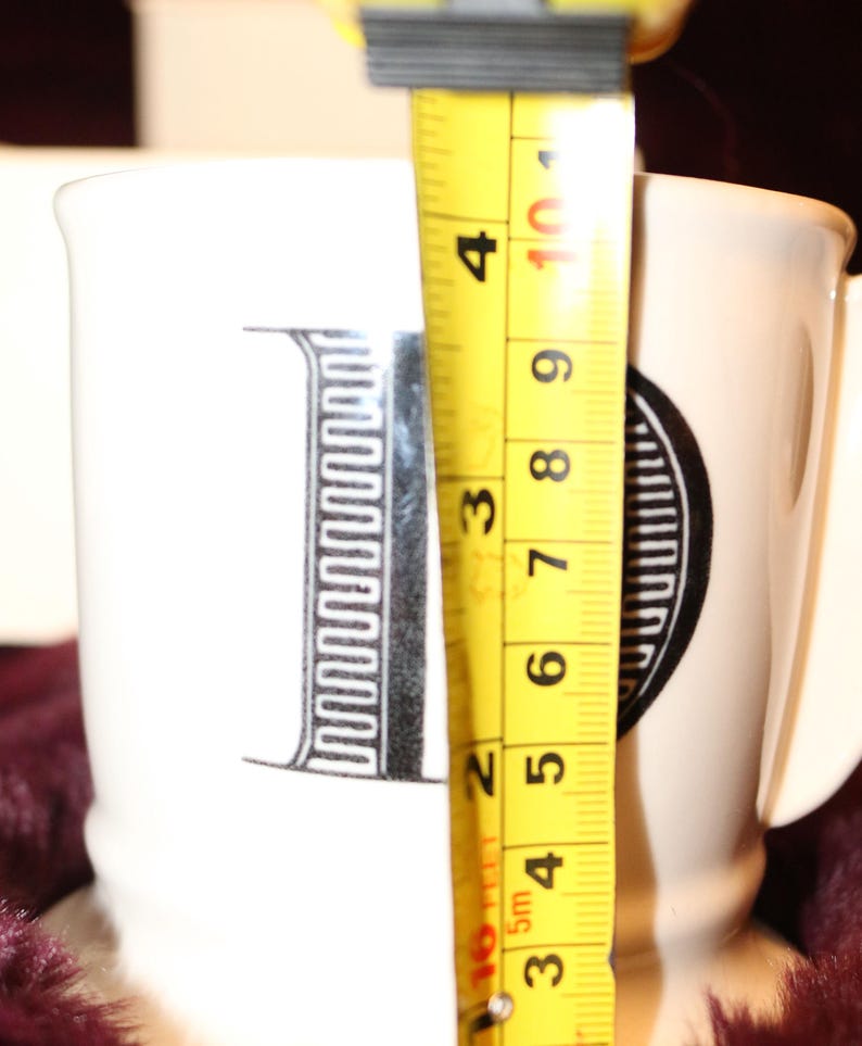 Pu&ograve; includere: Tazza in ceramica bianca con un design a lettera "I" nera. La tazza &egrave; alta circa 10 cm. La tazza ha un manico e si trova su una superficie viola scuro.