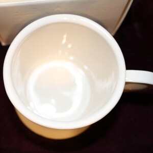 Pu&ograve; includere: Vista dall'alto di una tazza in ceramica bianca con manico. L'interno della tazza &egrave; di colore crema chiaro e il manico &egrave; bianco. La tazza &egrave; appoggiata su una superficie bordeaux scuro.