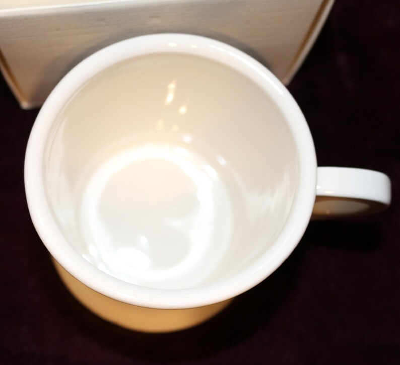 Pu&ograve; includere: Vista dall'alto di una tazza in ceramica bianca con manico. L'interno della tazza &egrave; di colore crema chiaro e il manico &egrave; bianco. La tazza &egrave; appoggiata su una superficie bordeaux scuro.
