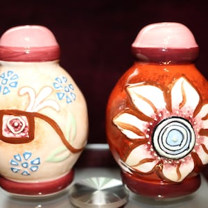 Könnte beinhalten: Ein Paar dekorative Salz- und Pfefferstreuer aus Keramik. Einer ist beige mit blauen Blumenakzenten und braunen Details. Der andere ist orange mit einem weißen Blumendesign. Beide haben rosa Deckel und weinrote Sockel.