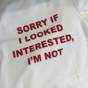 Op de afbeelding: Wit T-shirt met de rode tekst "SORRY IF I LOOKED INTERESTED, I'M NOT". Het shirt heeft een ronde hals en is gemaakt van een zacht materiaal. De tekst staat in het midden op de voorkant van het shirt.
