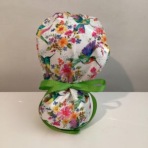 May include: A white scrub cap featuring a colorful pattern of hummingbirds and various flowers in shades of pink, yellow, and green. The cap includes a gathered back section secured with a bright green ribbon tied into a bow at the center.