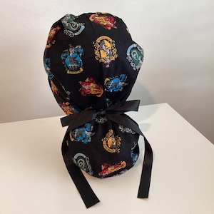 May include: Black scrub cap featuring colorful crests from a fantasy school. The cap has a black bow in the back for tying. The crests are in shades of red, yellow, blue, and green.