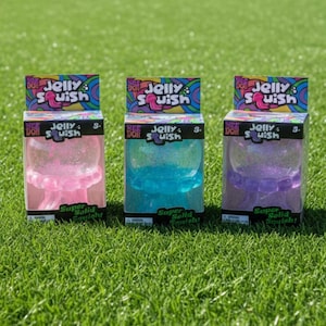 Può includere: Tre giocattoli Nee Doh Jelly Squish in scatole singole, con figure traslucide a forma di medusa di colore rosa, blu e viola con texture glitterate. Ogni scatola riporta il testo Jelly Squish e Super Solid Squish sulla confezione.