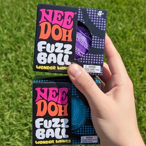 May include: Two square black boxes labelled Nee Doh Fuzz Ball Wonder Waves. The top box contains a purple textured ball, and the bottom box contains a blue textured ball. Each box features pink and orange text and a small circular cutout window.