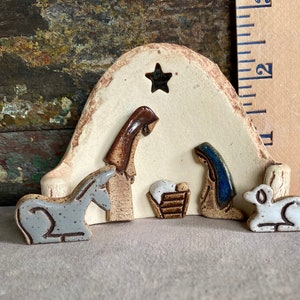 May include: A ceramic nativity scene with a white background and a star cutout. The figures include Mary, Joseph, a donkey, a lamb, and a manger with a baby Jesus.