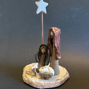 May include: A ceramic nativity scene figurine set on a circular base. It features two standing figures in brown and blue robes, a small figure in a white cloth in a brown manger, and a small sheep. A white star sits atop a thin copper wire rod.