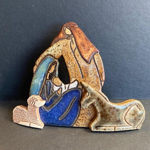 May include: A ceramic nativity scene featuring Mary, Joseph, and baby Jesus, with a lamb and donkey. The figures are in shades of brown, blue, and white, with a textured finish. The scene is a religious themed decoration.